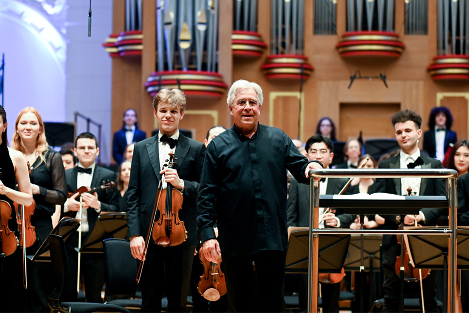 A male conductor, wearing smart black attire, standing next to the conductor's podium smiling at the audience, with musicians standing behind him.